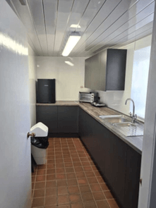 A narrow galley kitchen with dark grey cabinets, wood-effect countertops, and a terracotta tiled floor.