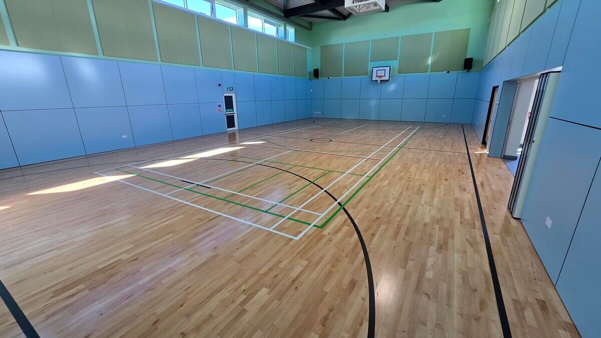 Bright indoor sports hall with polished wooden flooring, light blue paneled walls, multi-colored court lines, and a basketball hoop on the far wall.