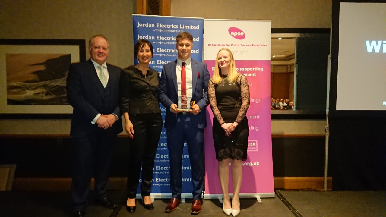 Four people standing in front of banners at the Scottish APSE annual awards event. One person is holding a glass award