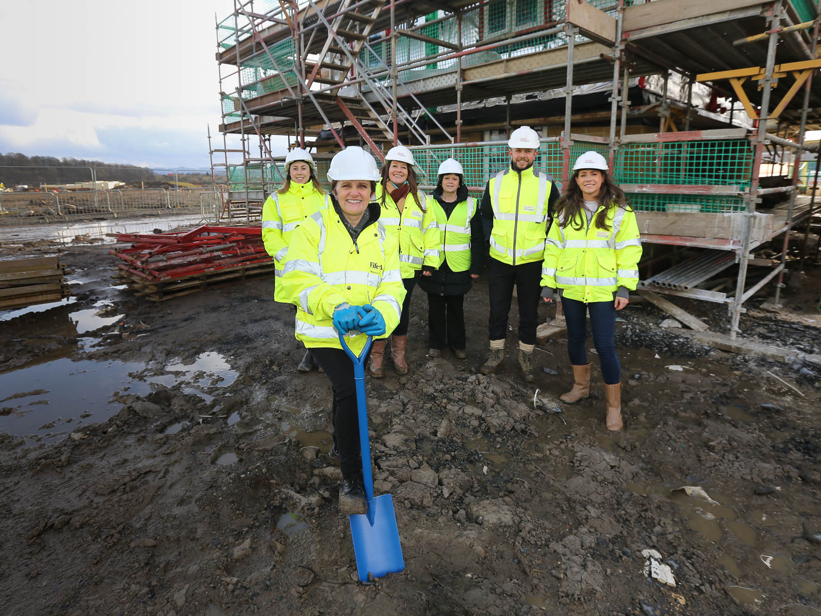 A group of people wearing high‑visibility jackets and hard hats stand together on a muddy construction site, with scaffolding and building materials in the background. One person in the foreground holds a blue shovel, positioned as if for a groundbreaking activity. - Work begins on new affordable homes for Inverkeithing