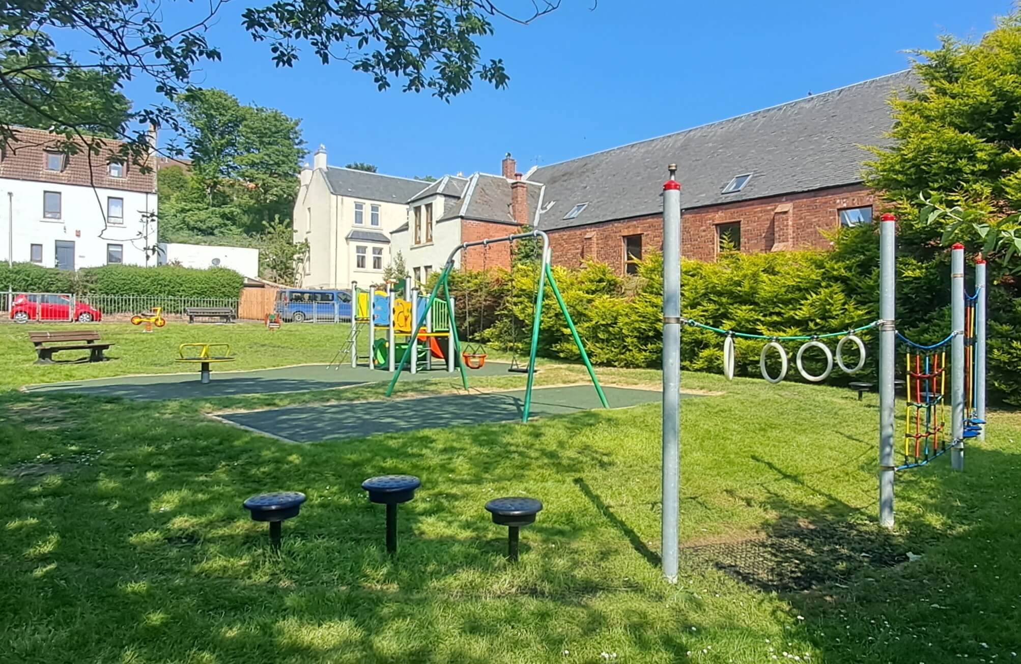 A playground with swings, slides, and climbing equipment. The playground is surrounded by green grass and houses in the background