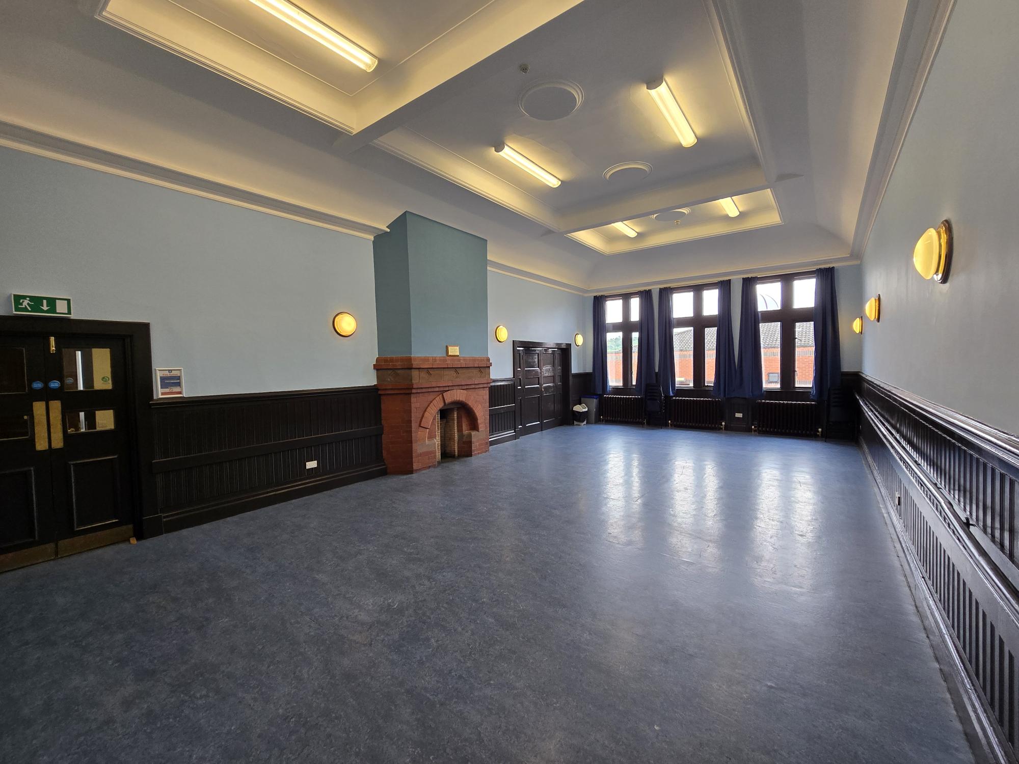 Empty room with blue walls, dark wood panelling, tall windows, and a small brick fireplace