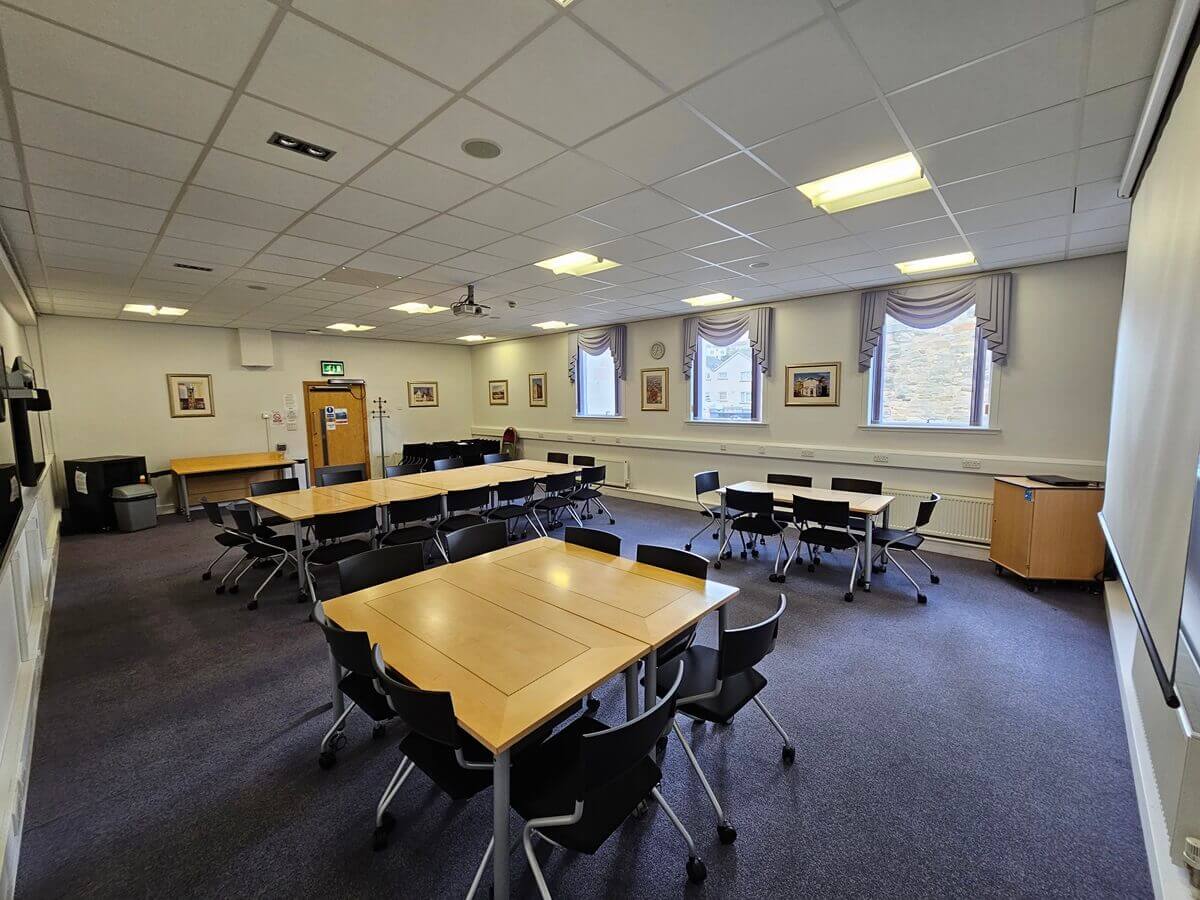 Large meeting room with several tables and chairs arranged in groups, bright overhead lighting, and three windows along one wall with framed pictures.