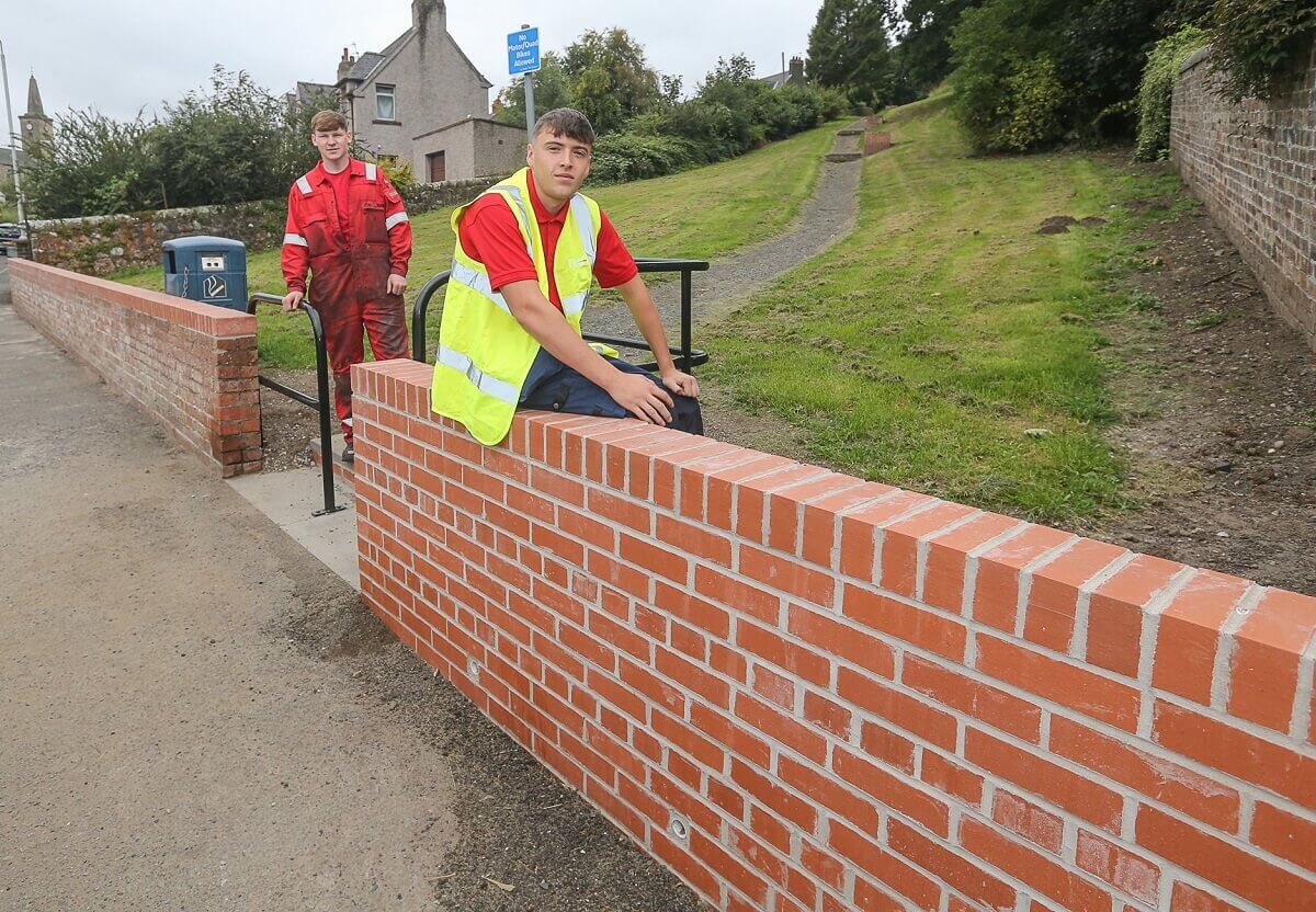Kyle Keddie, apprentice fabricator, stands next to railing he built and Harvey Ross, apprentice bricklayer sits on wall he built