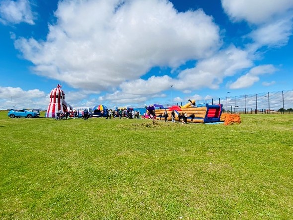 an outdoor event with a variety of inflatable structures on a grassy field under a blue sky with fluffy white clouds. There's a red and white striped inflatable slide resembling a lighthouse, a colourful inflatable dome, and other inflatable play structures. People are gathered near the inflatables. A blue car is parked on the left side of the image