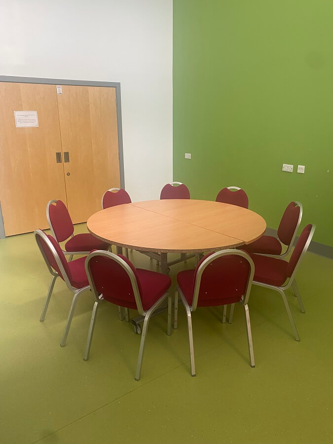 a room with a round table surrounded by red chairs. There are light wooden double doors on the left, and the walls are painted in white and light green.