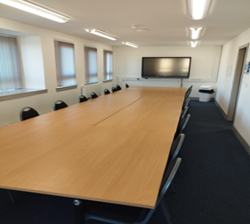 a meeting room with a long, light-colored table surrounded by black chairs. A large television screen is mounted on the white wall at one end of the table. The floor is covered with a dark blue carpet. There are windows with blinds on the left side of the room and a doorway on the right.