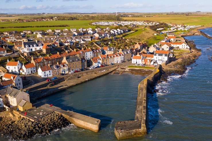 The image shows an aerial view of Cellardyke. The harbour is surrounded by stone walls extending into the sea, creating a sheltered area for boats. The village consists of numerous houses with red-tiled roofs and white walls, situated close to the coastline. There are green fields in the background and rocky shores along the water's edge.