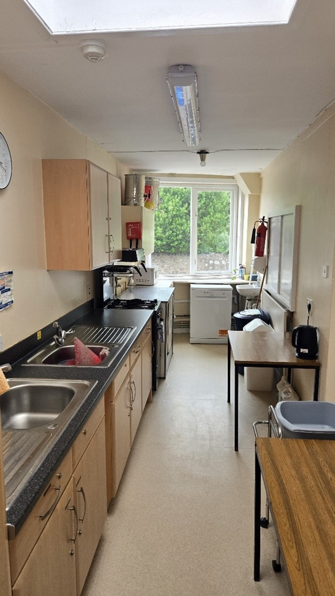 A narrow galley kitchen with light wood cabinets, black countertops, and two sinks on the left, leading toward a bright window at the end.