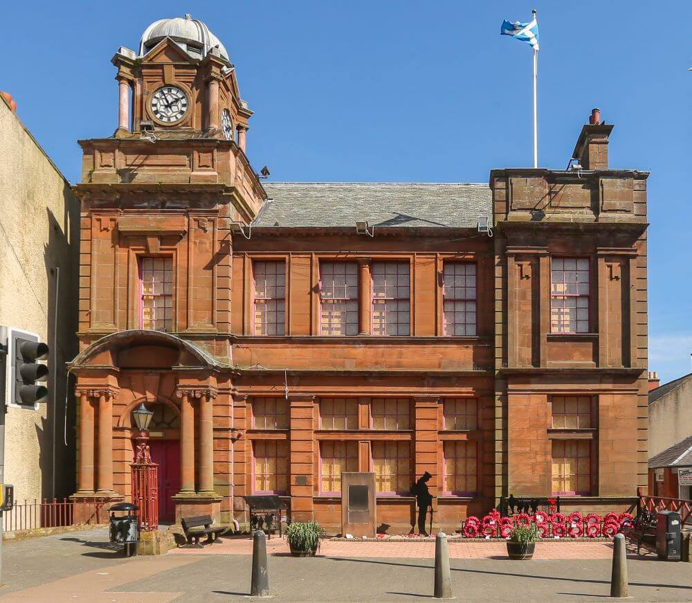 Front view of Cowdenbeath Town House, a red sandstone building with a clock tower and domed roof. A Scottish flag flies above the building, and a memorial with a silhouette figure and red poppy display stands in the paved area in front.