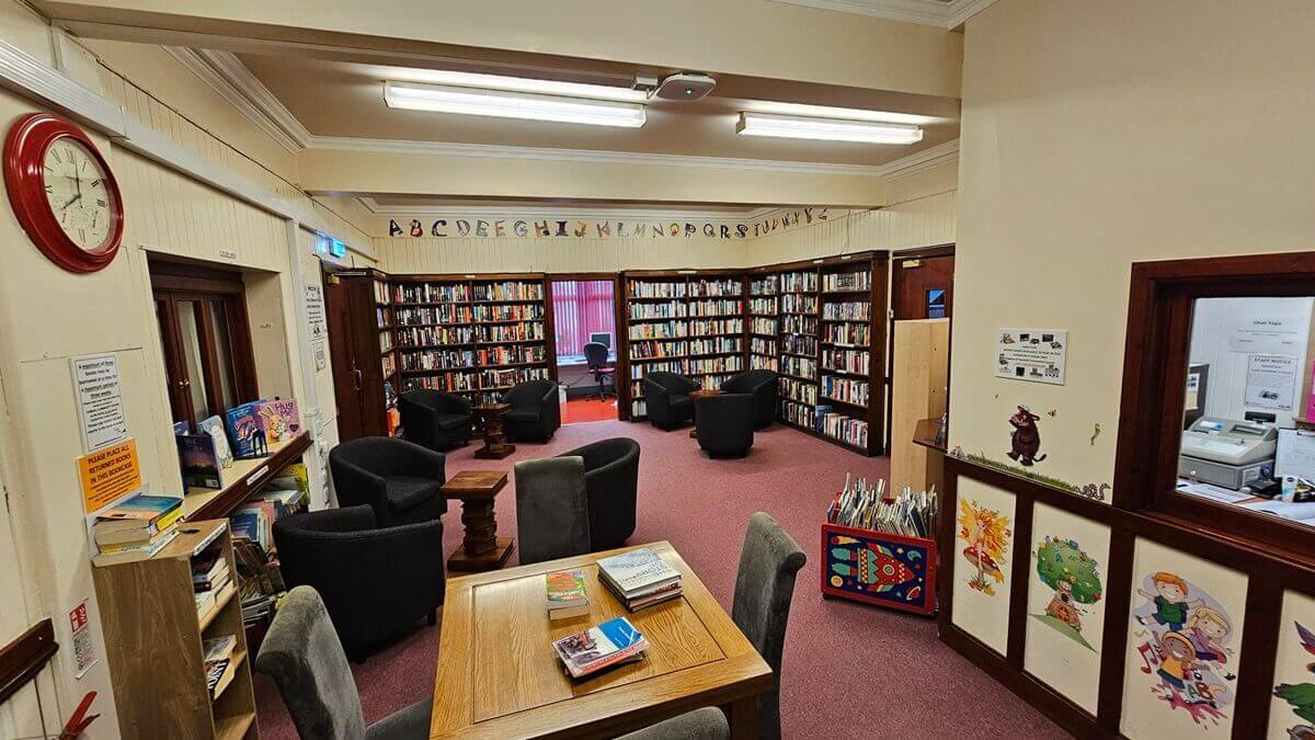 A cozy library room featuring dark wooden bookshelves, carpeted flooring, and several clusters of black armchairs for reading. A wooden study table sits in the foreground, and an alphabet banner stretches across the far wall.