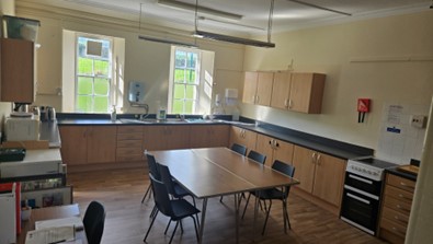 Kitchen inside a community hall with wooden cupboards, worktops, sink and appliances along the walls. A table with several chairs sits in the centre, and two windows provide natural light.