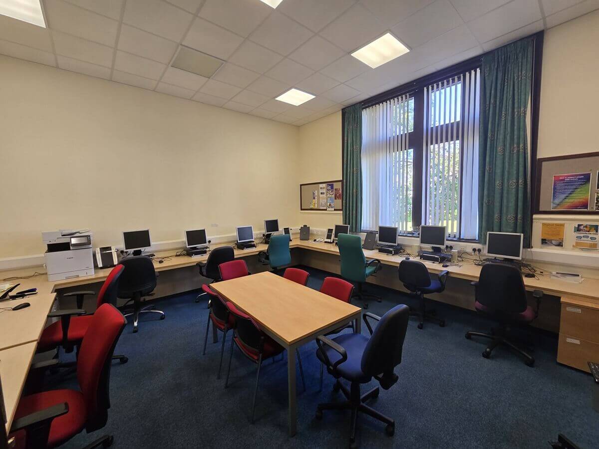 Computer room with desks along the walls, multiple monitors and chairs, a central table with chairs, and large curtained windows