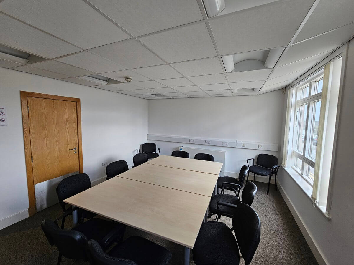Small meeting room with two tables pushed together, surrounded by black chairs, with a window on one side and a wooden door on the other.