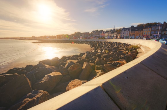 Kirkcaldy's sunny coastal promenade with curved sea wall, rocks, and buildings along the shoreline.