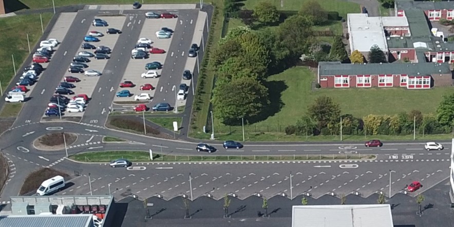 An aerial view of a parking lot filled with numerous cars. The parking lot is adjacent to a road with a roundabout and another smaller parking area. Green trees and buildings with red and white accents are visible in the background