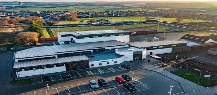 An aerial view shows a modern building complex with a silver roof, surrounded by a parking lot, sports fields, and green spaces
