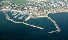 An aerial view of a Anstruther with a marina. The image shows two piers extending into the water, with numerous boats moored. Anstruther has a mix of residential and commercial buildings, green spaces, and roads leading to the waterfront. The coastline is curved, and the water appears calm.