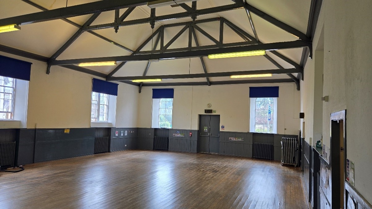 hall with polished wooden floors and an open timber-truss ceiling, featuring several large windows with blue blinds and grey wainscoting along the lower walls.