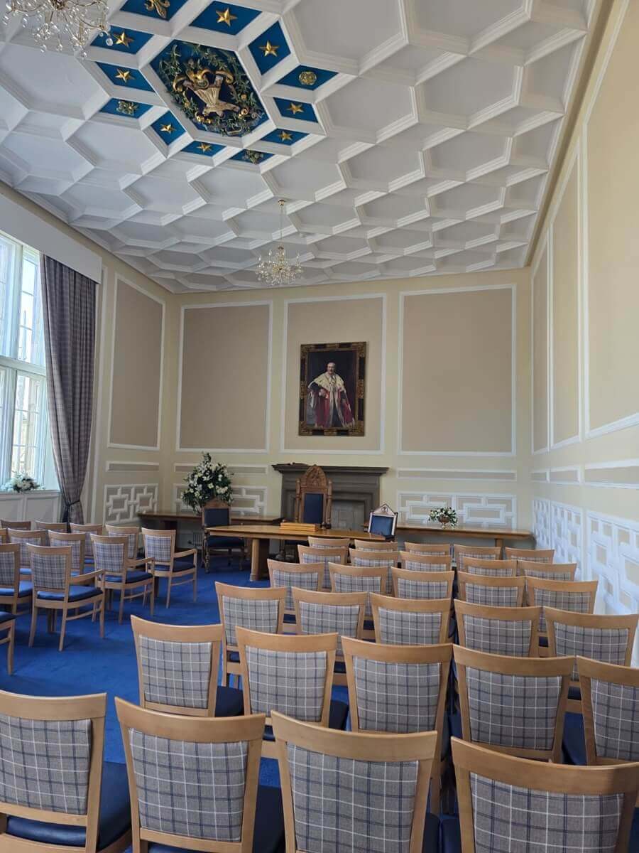 A grand ceremonial room featuring a blue carpet and rows of chairs with plaid backs facing a wooden desk. The high, white coffered ceiling includes a central blue and gold crest, and a formal portrait hangs above a fireplace in the background.