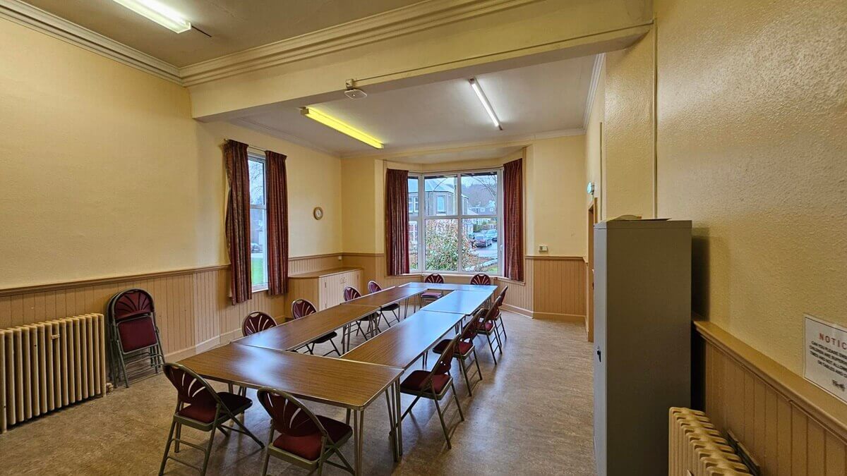 A brightly lit meeting room with pale yellow walls, wooden wainscoting, and a large bay window at the far end. Folding tables are arranged in a large U-shape in the center, surrounded by matching red-padded folding chairs.