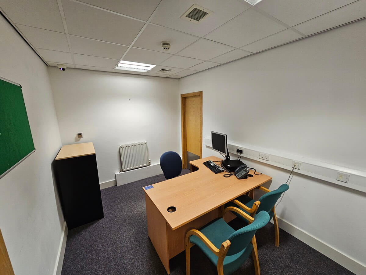 Small office with a wooden desk, computer, two visitor chairs, and a single office chair, with a door and radiator against the wall.