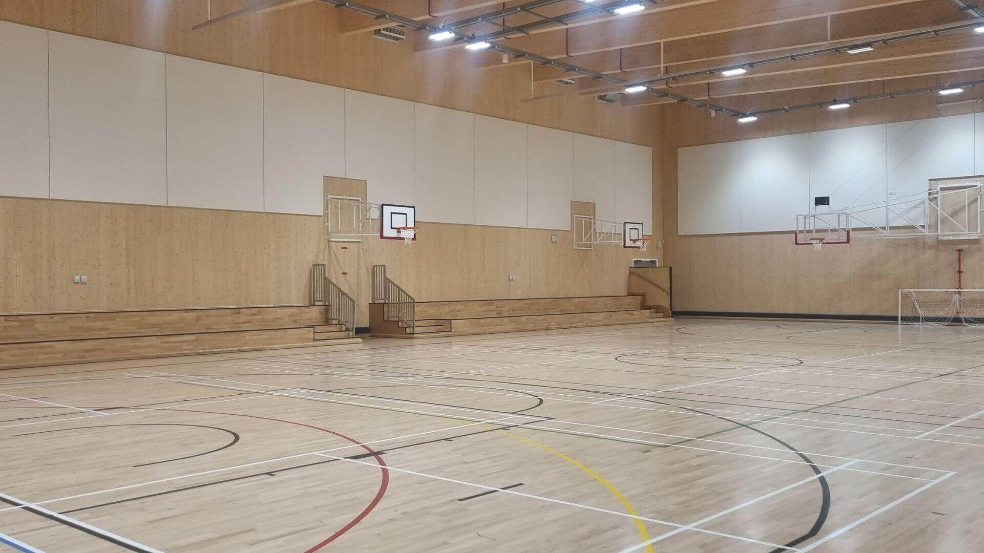 Multipurpose games hall with wooden flooring marked for different games such as indoor football and racket sports. Lots of white ceiling lights and several basketball nets on the wall.