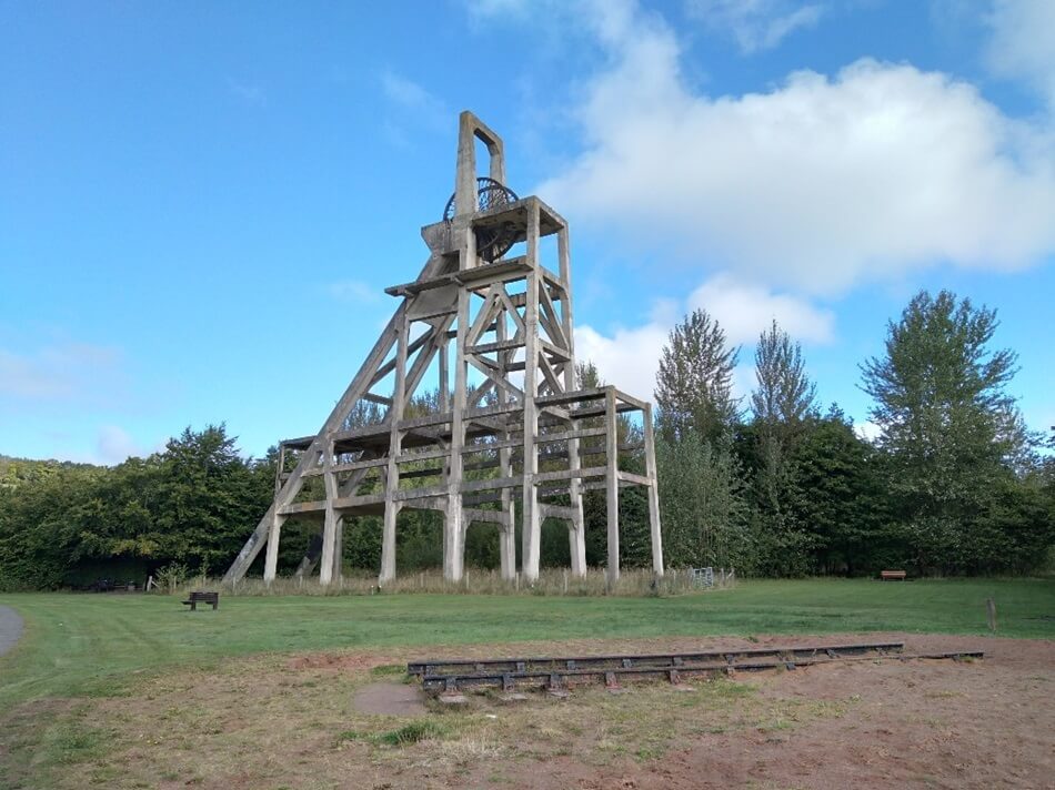 Historic mining structure with tall frame in a park, surrounded by trees and grass under a partly cloudy sky