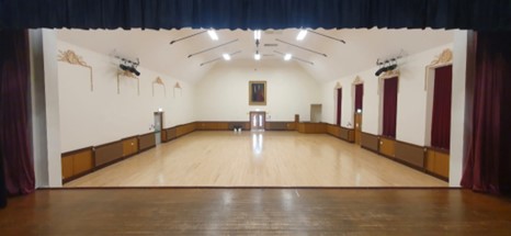 An empty, high-ceilinged assembly hall with a polished wood floor and red curtains, viewed from a stage