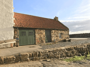 A small stone cottage with a red tiled roof and green doors, situated on a stone quay