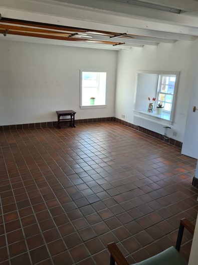 room with white walls and a reddish-brown tiled floor, featuring two windows with small potted plants on the sills and a white-beamed ceiling with several long wooden poles