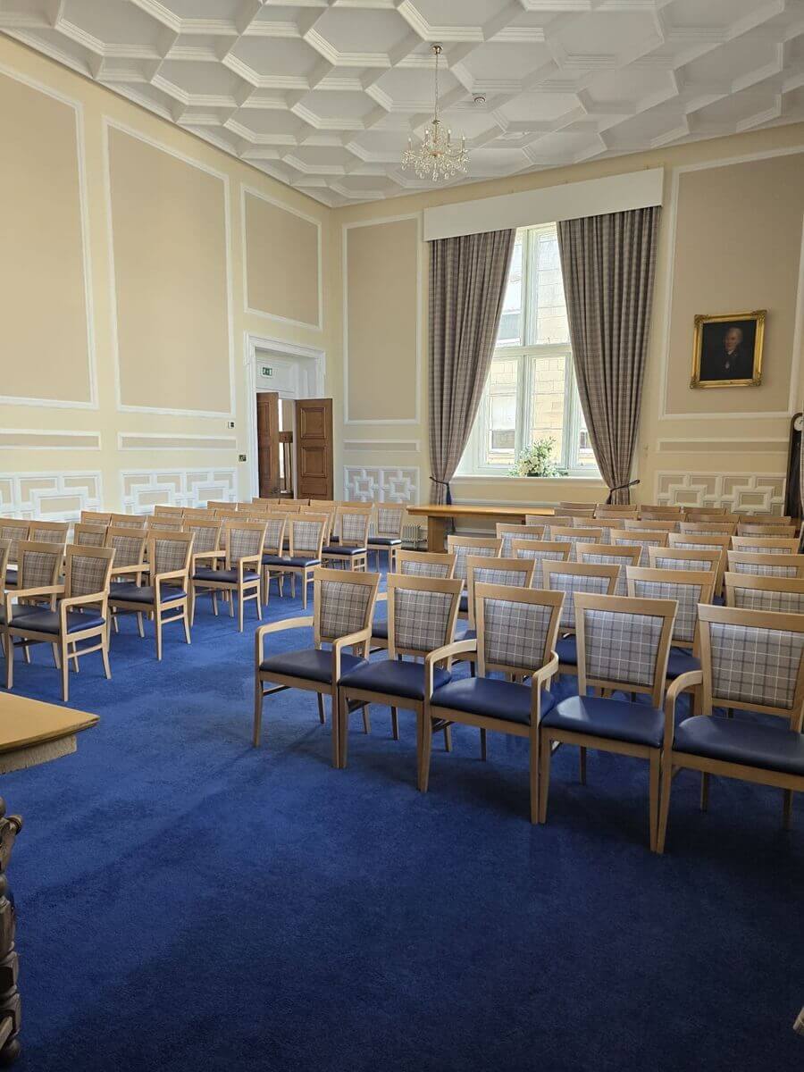 A large hall with dark polished wood floors and a high vaulted ceiling, featuring two bright windows in the center wall and chairs lined up along the perimeter.