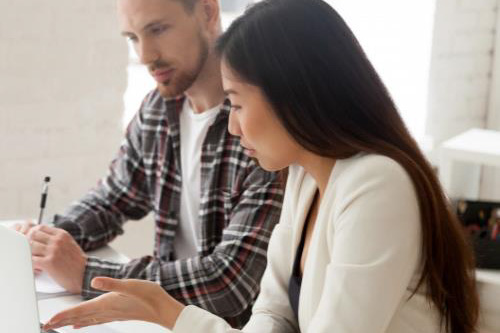 A man with checked shirt sitting next to a woman with a white top, talking and writing things down