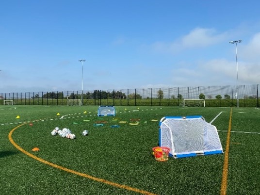 a green artificial turf football field under a blue sky. There are several footballs, small goals, and training markers scattered on the field. A black fence surrounds the field, and tall light posts are visible along the perimeter