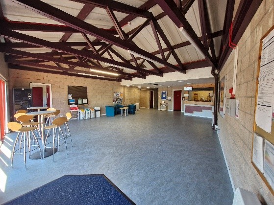 A spacious room with a high ceiling supported by exposed beams. There are several tables and chairs, including a tall round table with stools on the left side. The back wall has various informational materials and seating areas, while the right side features a counter area that appears to be for service or reception. There is also a bulletin board on the right wall with papers pinned to it. The room is well-lit with natural light coming through windows on the left side.
