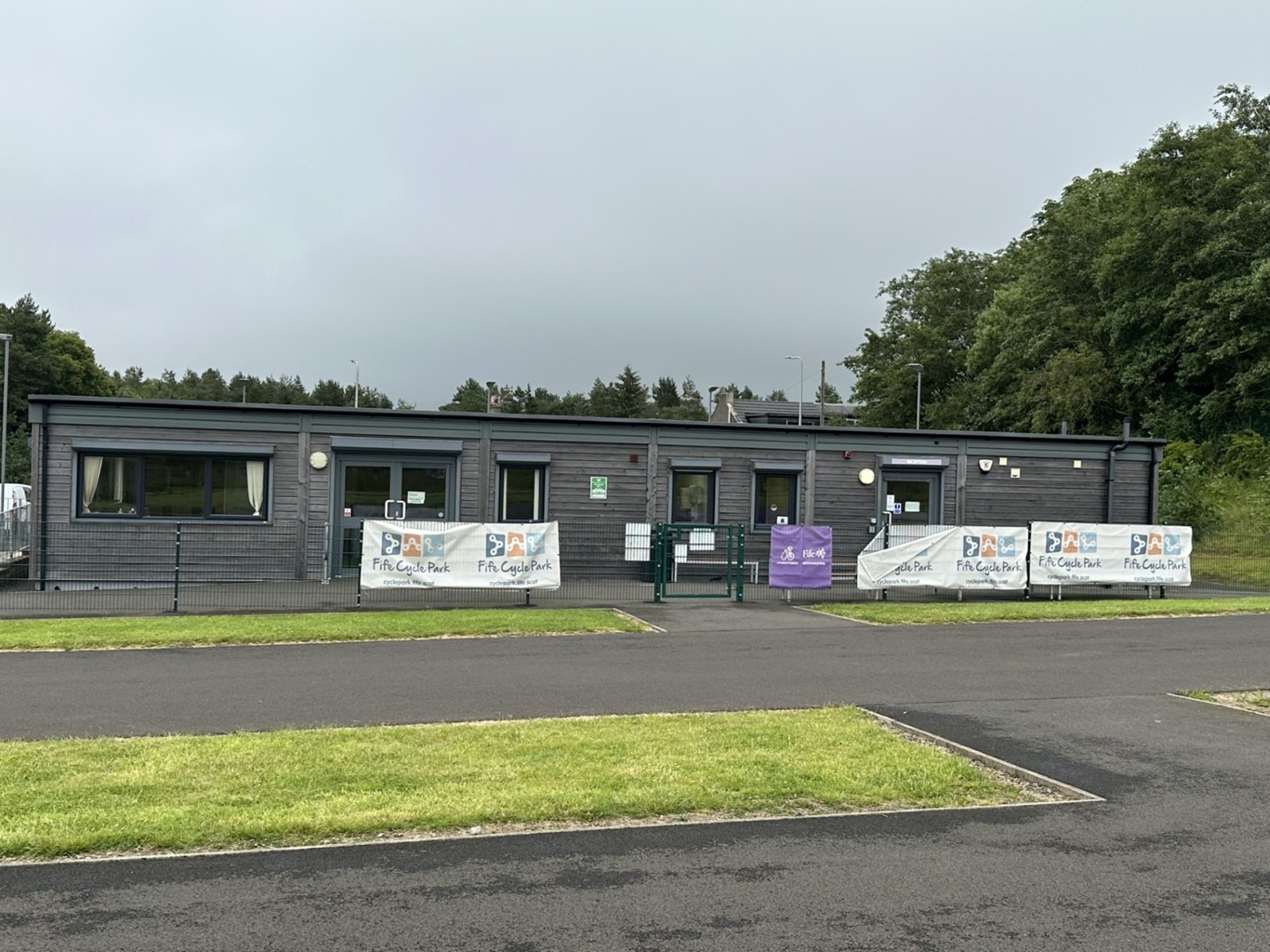 A single-story building with a grey exterior, surrounded by a paved area and patches of grass. There are several banners displayed on the fence in front of the building, with text that reads 'Fife Cycle Park' and images of cyclists. There is also a purple banner near the entrance with text that is not fully visible. The background includes trees and some distant structures.