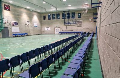three rows of empty blue chairs lines the right side of a school gymnasium with a green floor, facing a basketball court with hoops in the distance