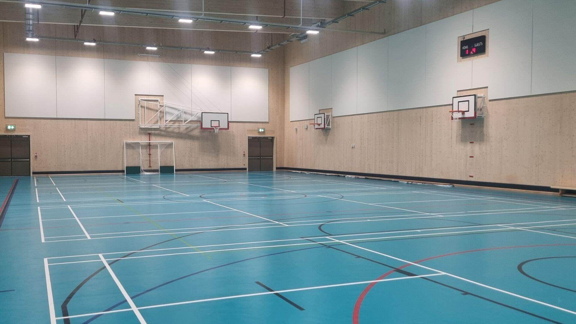 Multipurpose hall with blue floor and markings for different net sports. Wall is painted white and brown and has several basketball nets on the wall. There are lots of white ceiling lights above and a timer on the right wall.