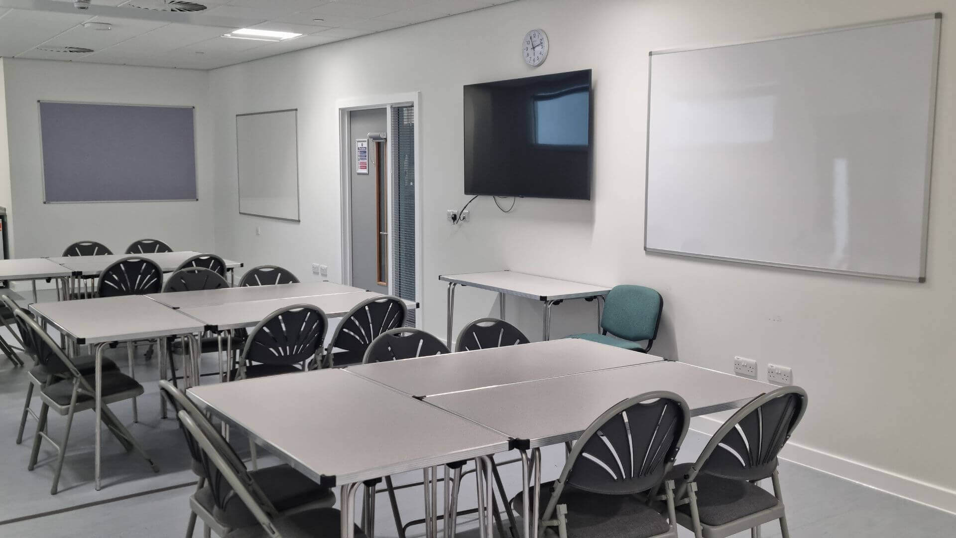Brightly lit meeting room three tables, each surrounded with five chairs. The room has a white smart boards and a television hung on the wall,. There is an extra table with a green chair in front of the room