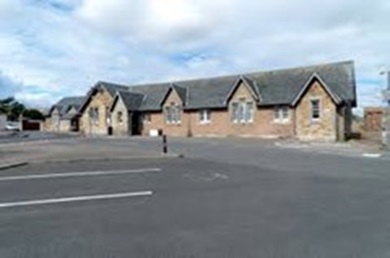 A long, single-story stone building with a dark pitched roof and several gabled windows, situated behind an asphalt parking lot