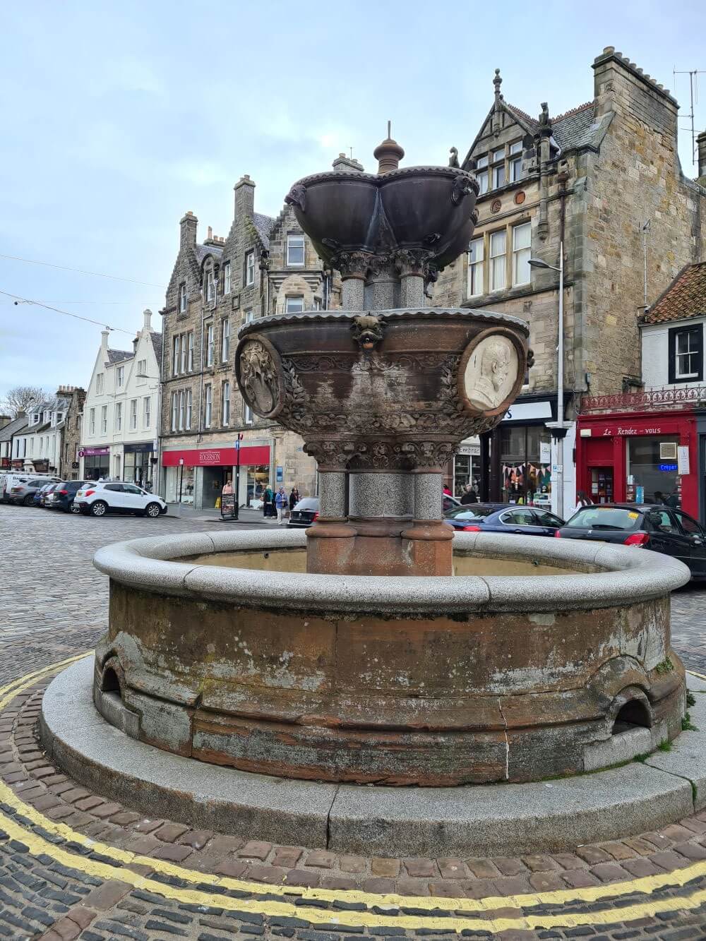 A stone fountain stands in the centre of a cobbled town square, surrounded by parked cars and traditional stone buildings with shops at street level.