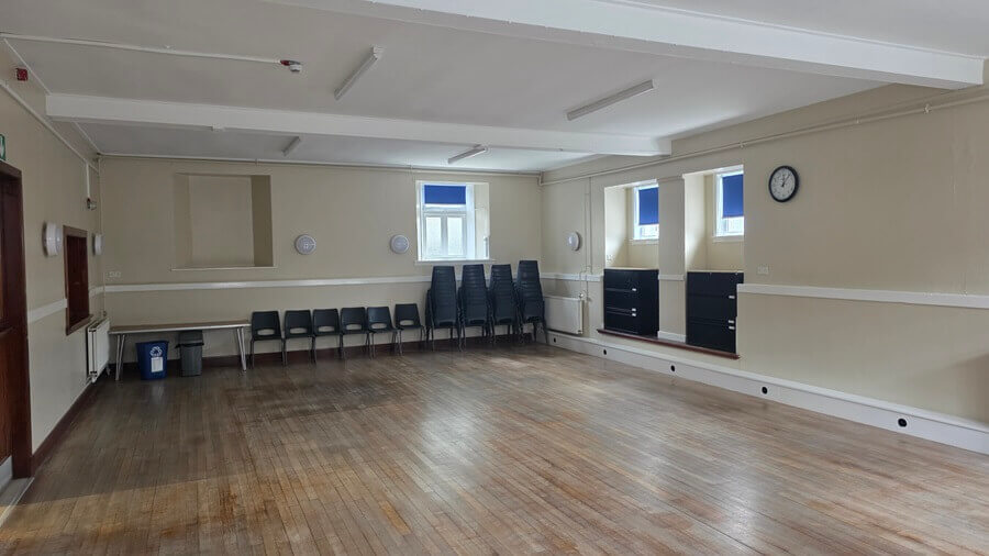 An empty community hall with a wooden floor, beige walls, and dark plastic chairs lined up and stacked against the back wall.