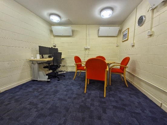 A small meeting room with blue carpeted flooring and cream-coloured walls. There are four orange chairs around a wooden table in the center of the room. On the left side, there is a desk with a computer monitor, keyboard, mouse, and an office chair. The walls have some electrical outlets and two white rectangular devices mounted on them. A clock is visible on the right wall along with a framed picture. The ceiling has two circular light fixtures providing illumination to the room.