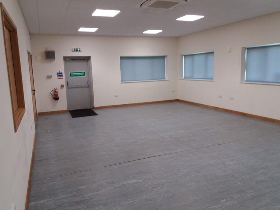 An empty, plain room. It has a gray floor. Windows with blinds line one wall.