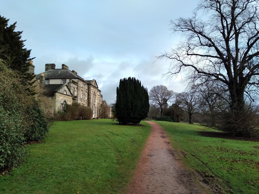 Oswald House, a historic stone mansion in Dunnikier Park, with a gravel path, green lawn and surrounding trees under an overcast sky