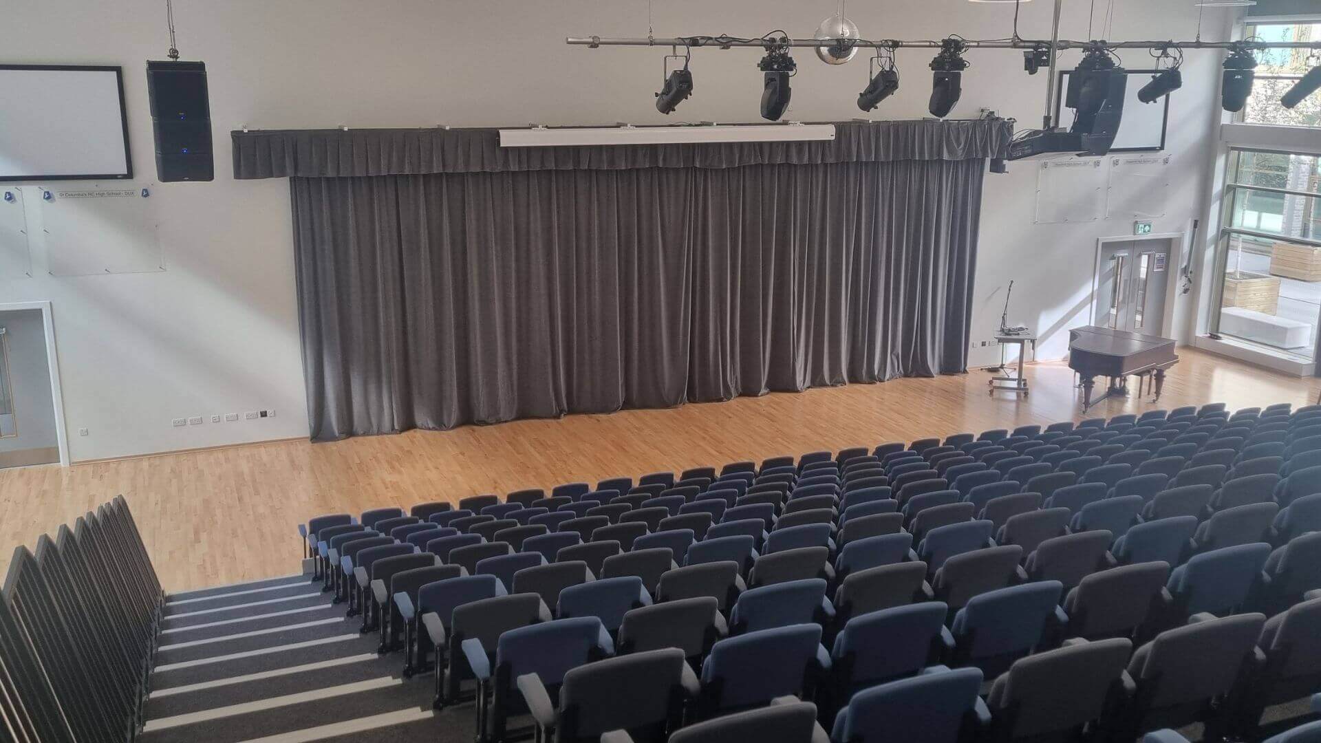 Large Hall with descending seats, the stage has brown wooded floor and grey curtains. The stage also has several lights hanging above it, a table and large window to the far right 