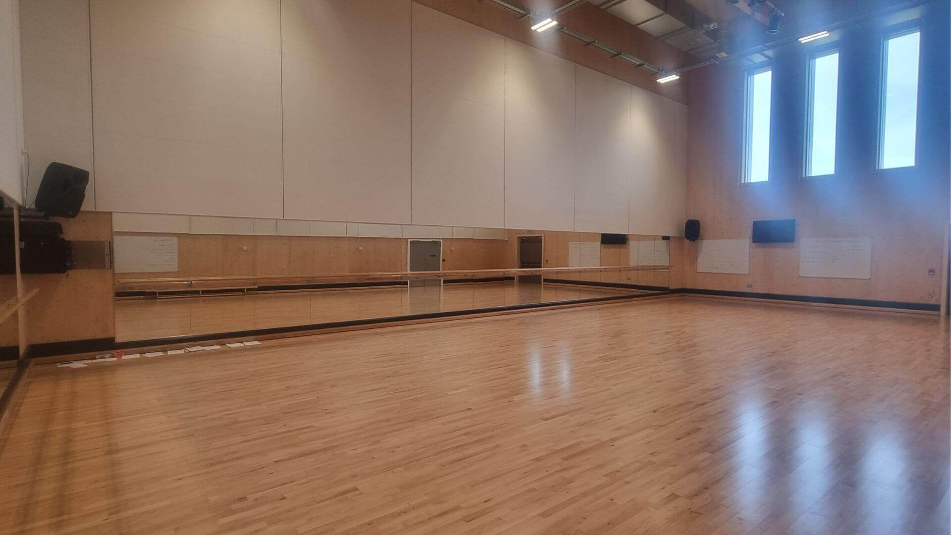 Large bare hall with wooden floors, large windows, large mirrors on the white walls. there are several black speakers hung on the wall and white lights on the ceiling