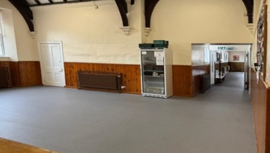 An open indoor space with grey flooring and wood-panelled lower walls, featuring high arched ceilings with dark exposed beams and a hallway leading off to the right.