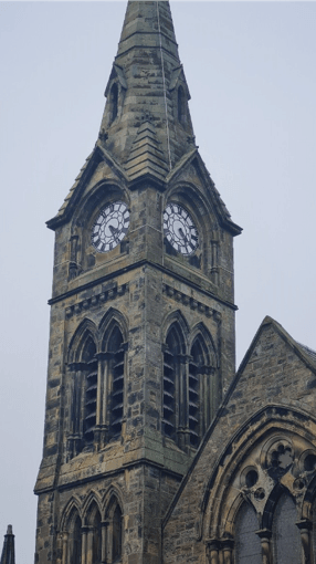 A close-up shot of a stone Gothic-style church tower featuring arched louvered windows, two prominent clock faces, and a pointed spire extending upward against a gray sky.