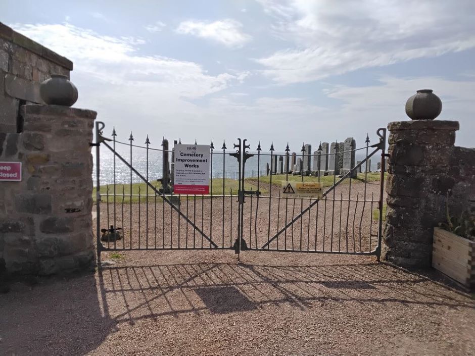 Closed metal gates at the entrance to St Monans Cemetery, flanked by stone pillars. Warning signs are attached to the gates, including one about cemetery improvement works. Beyond the gates, rows of gravestones stand on a grassed area overlooking the sea under a bright, partly cloudy sky. - Headstone inspections at St Monans Churchyard & Cemetery
